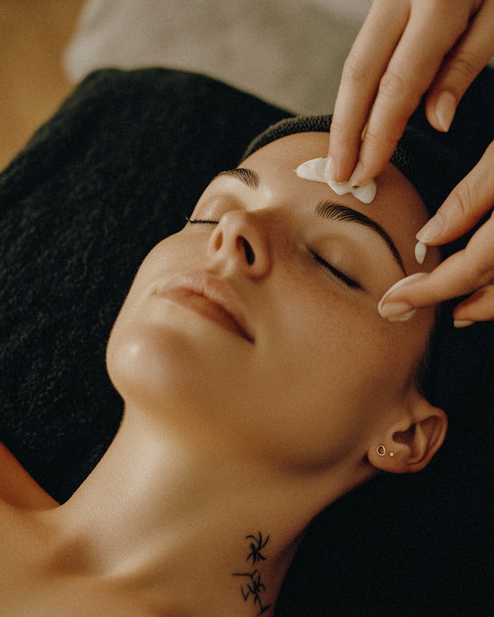 A woman with a neck tattoo relaxes with closed eyes during a facial beauty treatment, infused with warm ambiance.